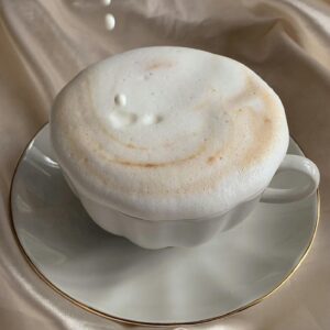 Delicate cappuccino topped with foam as milk pours, set against luxurious satin fabric.
