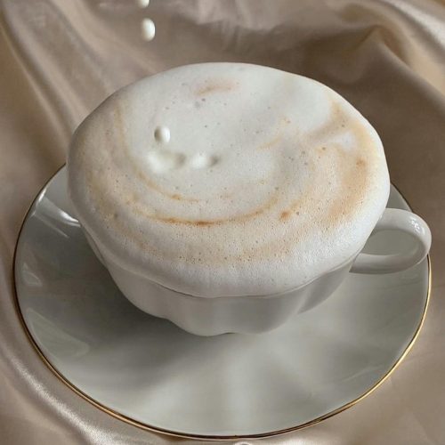 Delicate cappuccino topped with foam as milk pours, set against luxurious satin fabric.