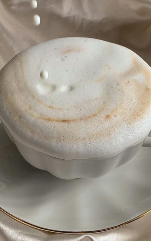 Delicate cappuccino topped with foam as milk pours, set against luxurious satin fabric.
